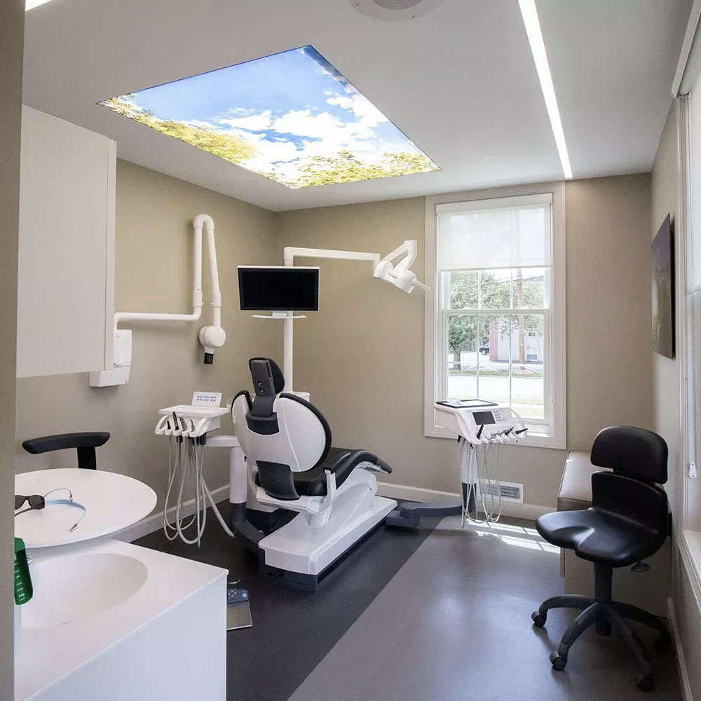 The image shows an interior view of a dental office with modern equipment, including a dental chair, examination tables, and various dental tools, set against a backdrop of a beige wall with a large skylight allowing natural light into the space.